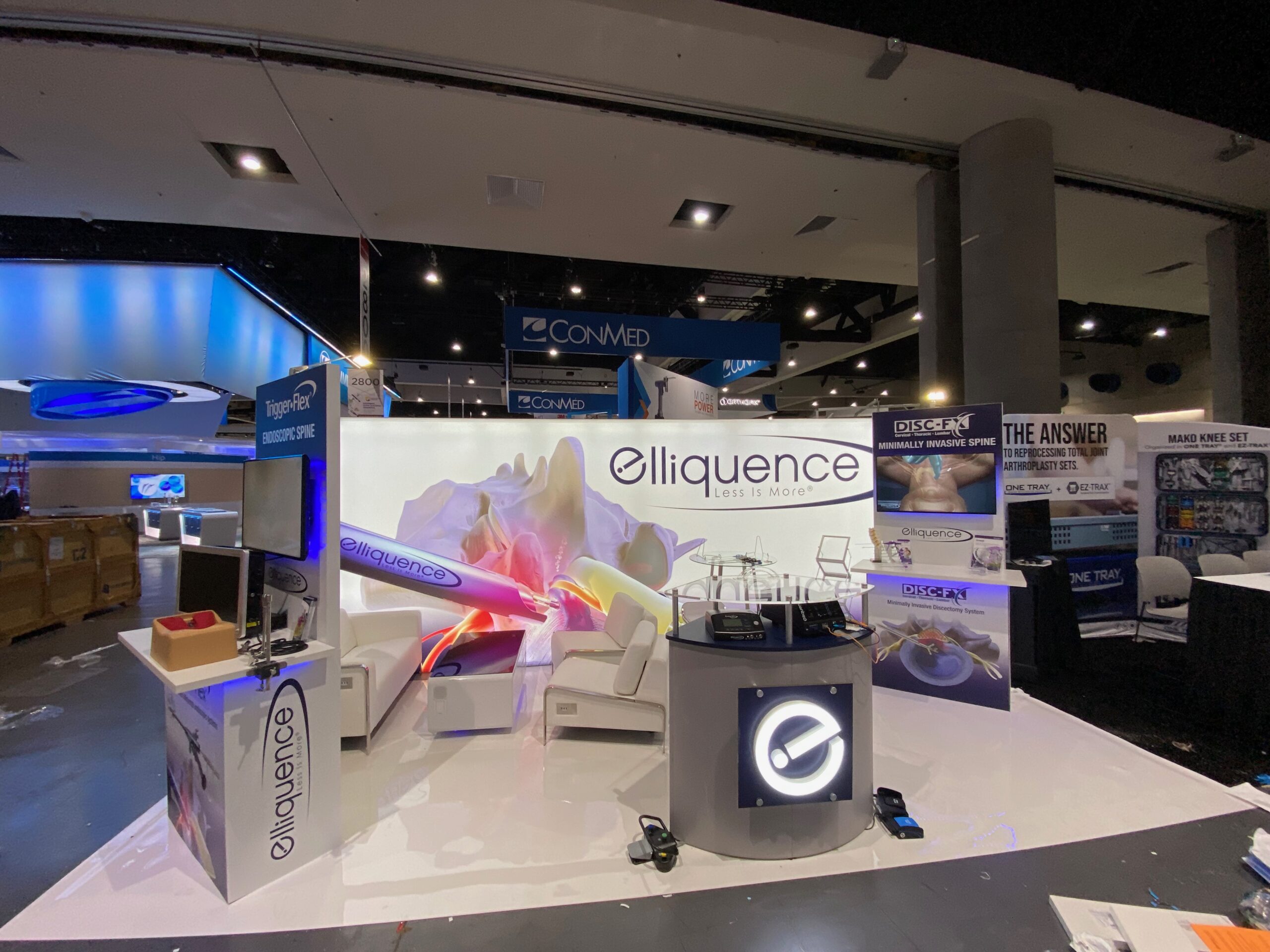 10x20 custom trade show booth featuring product display counters, multiple TVs, a light box backwall with custom company graphics, illuminated company logo, and a modern lounge area for attendees.