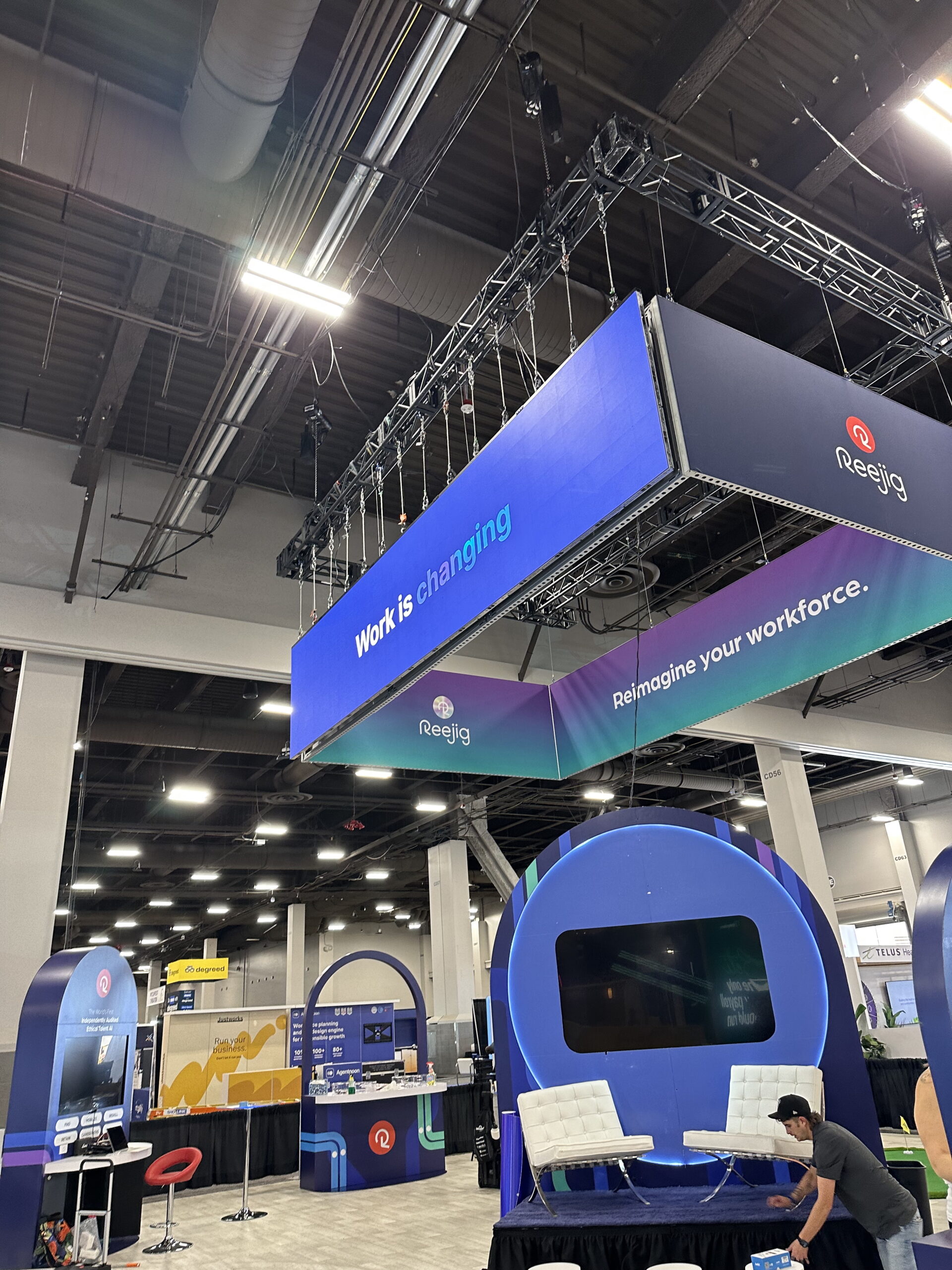 Reejig trade show booth featuring a rigged LED hanging sign suspended from the ceiling, constructed with seamless LED tiles for high visibility. The vibrant display reads “Work is changing” and “Reimagine your workforce,” enhancing brand presence in the exhibition hall. The booth includes modern seating and digital displays, creating an engaging environment for visitors.