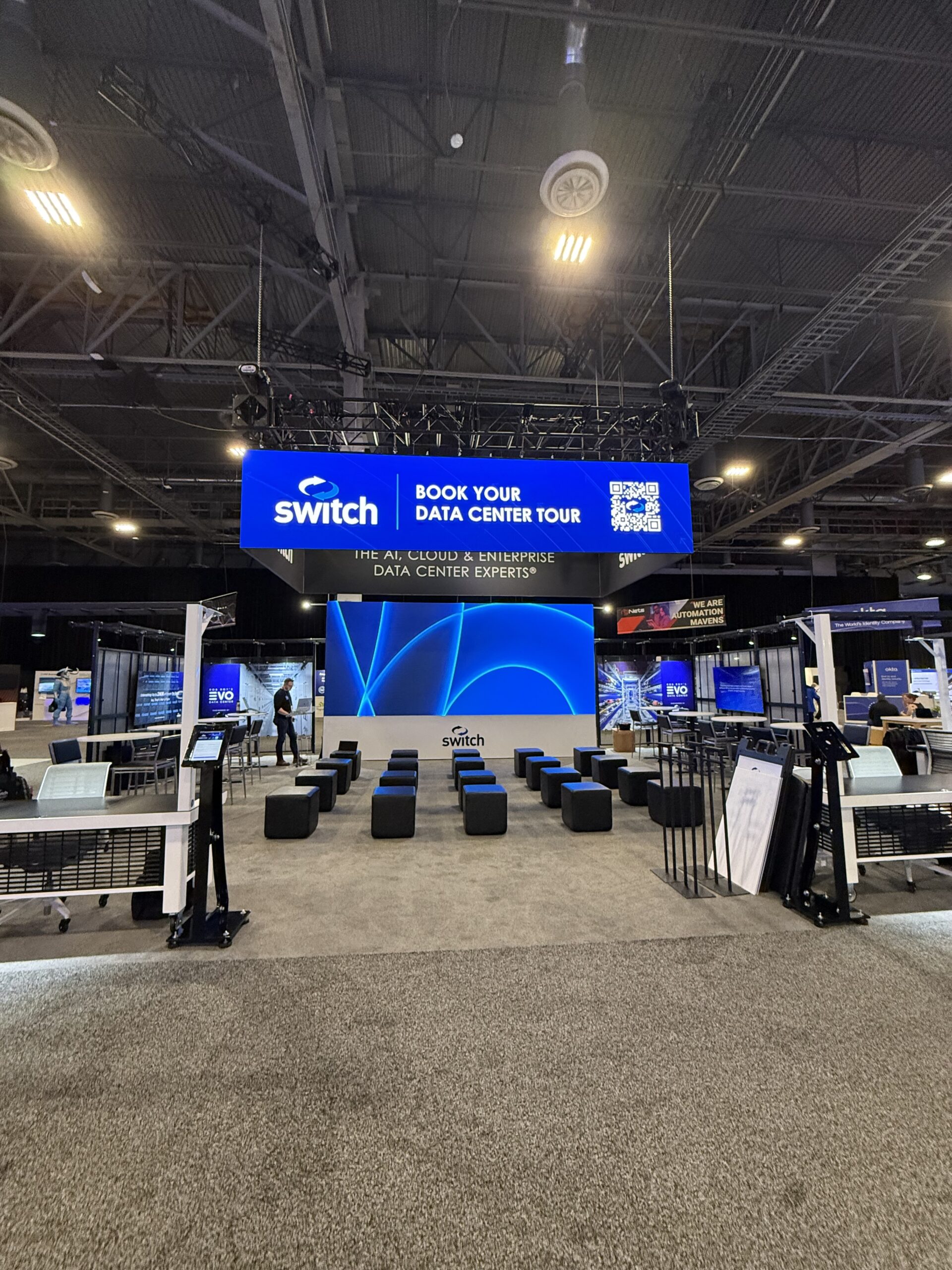 A trade show booth for Switch, a data center services company, featuring a modern setup with a large LED wall displaying a blue abstract design. Above the booth is a rigged LED hanging sign with the Switch logo and the text “BOOK YOUR DATA CENTER TOUR” alongside a QR code. The booth includes a theater-like seating arrangement with rows of black cube seats facing the LED wall, several kiosks with screens, and informational displays.