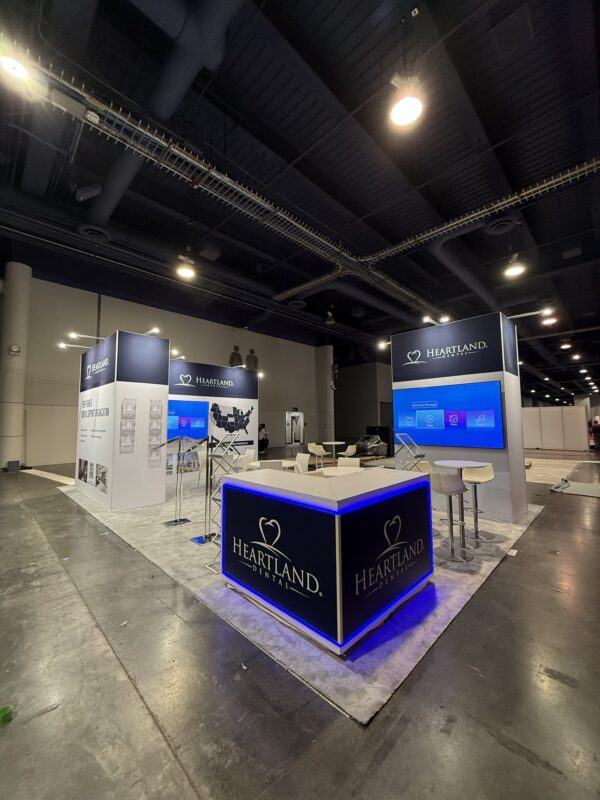 Heartland Dental booth at the ICSC trade show featuring illuminated counters, custom client graphics, wall-mounted TVs, literature racks, and seating areas on a carpeted floor.