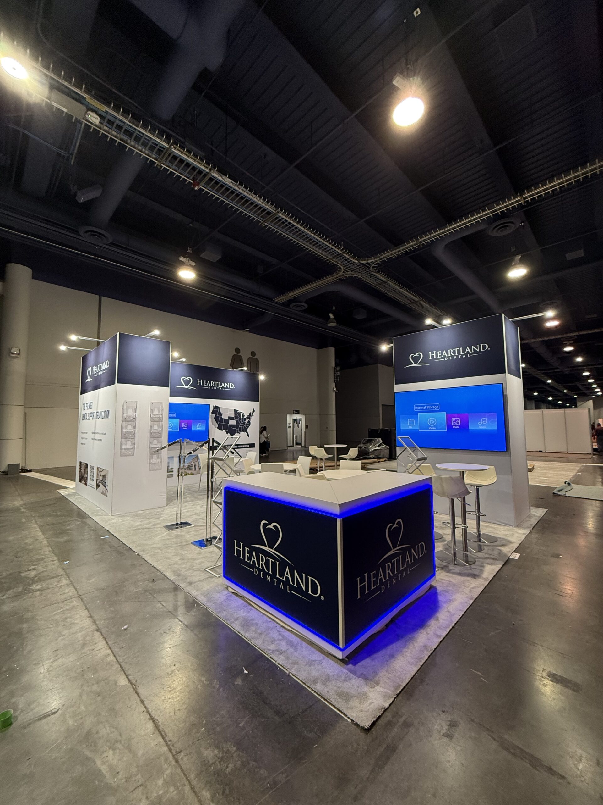 Heartland Dental booth at the ICSC trade show featuring illuminated counters, custom client graphics, wall-mounted TVs, literature racks, and seating areas on a carpeted floor.