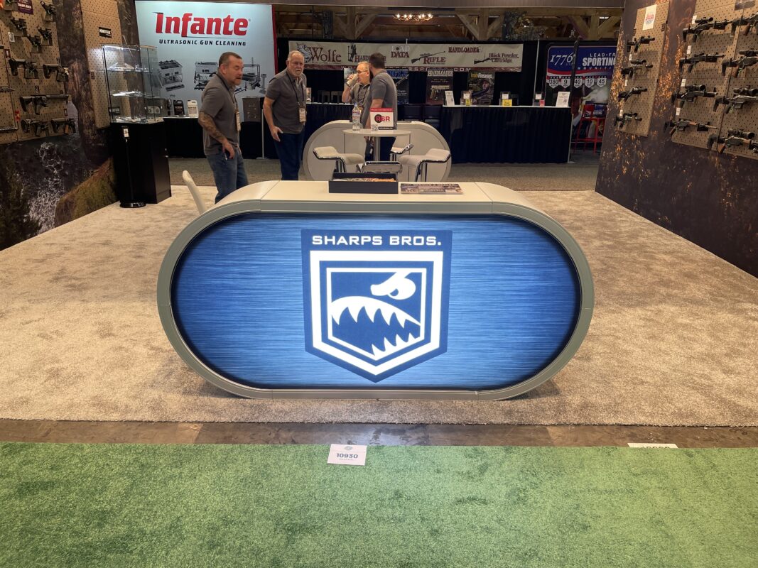 Custom backlit pill counter for Sharps Bros with an illuminated shark logo on a brushed metal face, surrounded by firearm displays and branded graphics at the SHOT Show in Las Vegas.